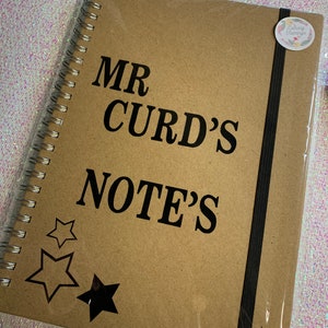May include: A brown spiral notebook with a black elastic closure. The cover has the text "MR CURD'S NOTE'S" in black lettering and three black star shapes.
