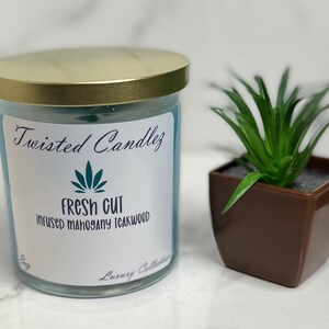 Könnte beinhalten: Ein tealblaues Kerzengefäß mit einem goldenen Deckel. Das Etikett lautet "Twisted CandleZ" und "Fresh Cut Infused Mahogany Teakwood".