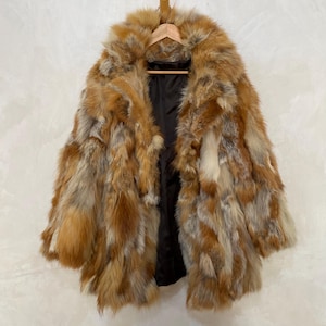 Può includere: Un lussuoso cappotto di pelliccia lungo in tonalità marrone, beige e grigio, appeso a una gruccia di legno. Il cappotto presenta un ampio colletto e una fodera scura, mostrando un design classico. La pelliccia appare morbida e soffice.