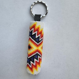 Peut inclure: Un porte-clés perlé avec un design cylindrique. Le porte-clés présente un motif de perles blanches, orange, jaunes et bleues. Le haut est doté d'un porte-clés argenté et d'une lanière en cuir noir.