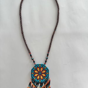 Peut inclure: Un collier de perles avec une chaîne en forme de corde marron. Le pendentif est un octogone turquoise avec un motif central en forme d'étoile en rouge, jaune et orange. Le pendentif a une frange de perles noires, oranges et turquoise.