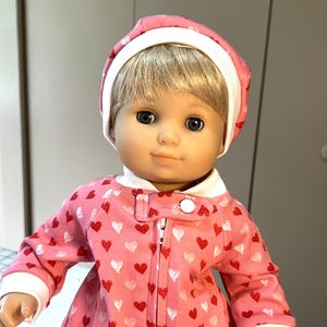 May include: A doll wearing a pink and white heart-patterned dress and a pink and white hat. The doll has blonde hair and blue eyes.