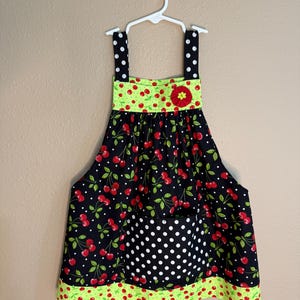 May include: A black and red cherry print apron with a white polka dot pocket and green trim. The apron has black straps with white polka dots and a green band with red cherries. A red flower is on the front.