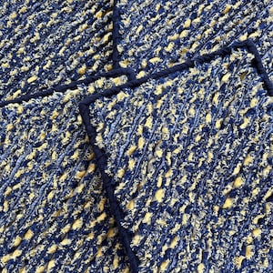 May include: Close-up of several square, textured placemats. The placemats are primarily navy blue with yellow accents, and a dark blue border. The fabric has a woven appearance, creating a tactile and visually interesting surface.