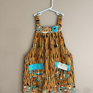 May include: A children's orange and black tiger stripe print dress with turquoise accents. The dress has two pockets with a tiger face print and a turquoise flower detail. The dress is hanging on a white hanger.