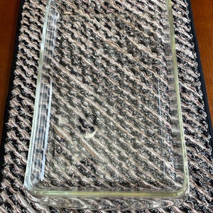 May include: A clear rectangular glass baking dish. The dish is empty and sits on a black, white, and brown patterned placemat. The placemat has a black border and is on a wooden surface.