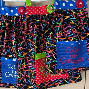 May include: Three colorful aprons with a crayon print and pockets. The pockets have embroidered text that says "Color me Creative", "Color me Creative", and "Color Out of the Box".
