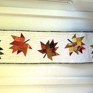 May include: A rectangular quilted table runner featuring a white background with five appliqued maple leaves. Each leaf is constructed from a patchwork of various fabric squares in shades of green, orange, red, yellow, and brown.
