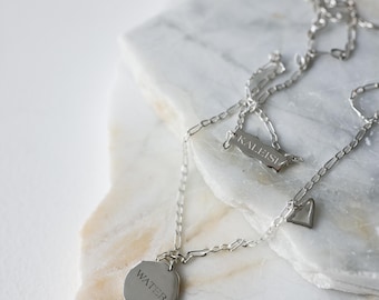 May include: A silver necklace with multiple charms, including a heart and a geometric shape with the word "WATER" engraved on it. The necklace is displayed on a white and gray marble surface.