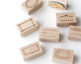 May include: A collection of wooden stamps with various engraved designs, including an oval, a rectangle, a book, and a gemstone. Each stamp is rectangular and features the text "NYRET DESIGN". The stamps are light brown in color.