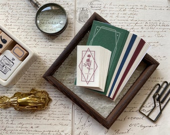 Puede incluir: Una pila de tarjetas en blanco con un diseño floral en la tarjeta superior, expuesta en un marco de madera oscura. Las tarjetas están dispuestas en un arcoíris de colores, incluyendo verde, azul, rojo y blanco.