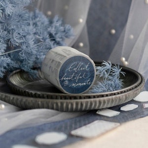 May include: A roll of decorative washi tape with the words "Collect beautiful moments" in white script on a dark blue background. The tape is on a grey metal plate with blue floral accents and a sheer fabric backdrop.
