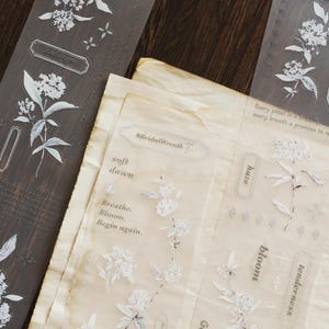 May include: Floral washi tape and stickers featuring white flower illustrations. The tape is dark brown and the stickers are on translucent paper. Text includes "#BridalWreath", "soft dawn", "Breathe. Bloom. Begin again."