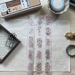 May include: Washi tape with a floral rose pattern in pink and purple hues. The semi-transparent tape is suitable for crafts and journaling. Other stationery items, such as stamps and a magnifying glass, are also visible.