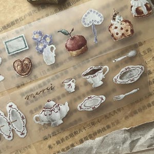 May include: A clear sticker sheet featuring various watercolor-style illustrations including a teapot, teacups, plates, a fork, a spoon, pastries, a small pitcher, a vase with purple flowers, and the handwritten word merci in cursive script.