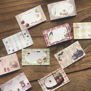 May include: A collection of ten rectangular paper memo pads with scalloped edges, each featuring unique watercolor-style illustrations including food, a cat, flowers, and text like Bon appétit and Have a nice day, arranged in a scattered layout.