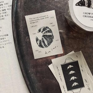 Könnte beinhalten: Zwei rechteckige Papieraufkleber mit schwarzer Tinte. Ein Aufkleber zeigt ein Croissant mit dem Text 'The curved croissant is traditionally made with margarine.' Der andere zeigt ein Backblech mit vier Croissants und '180°C | 18-20 min'.