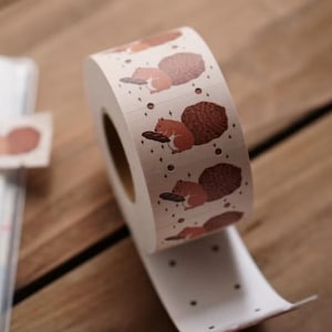 Op de afbeelding: Een rol decoratieve washi-tape met een herhalend patroon van cartoon eekhoorns die eikels vasthouden. De tape is gebroken wit met bruine en oranje illustraties en kleine zwarte stippen. De tape ligt op een houten oppervlak.