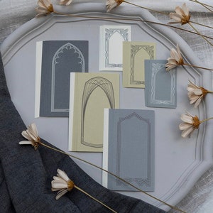May include: Six decorative paper frames with an arched design. The frames are in shades of grey and beige. The frames are on a white surface.