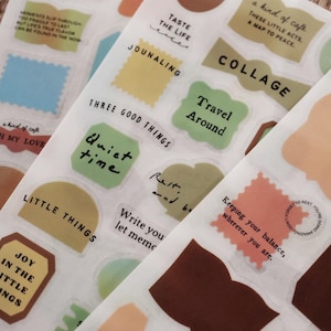 May include: A sheet of clear adhesive stickers featuring various shapes in muted tones of green, yellow, brown, and blue. The stickers include printed text such as TASTE THE LIFE, COLLAGE, Travel Around, Quiet time, LITTLE THINGS, and JOY IN THE LITTLE THINGS.