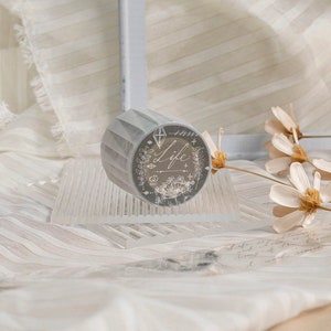 May include: A decorative washi tape roll with a grey geometric design. The tape features the word "Life" in elegant script, surrounded by floral and botanical illustrations. The tape is displayed on a clear, textured surface.