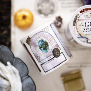May include: A roll of decorative tape with a gem design and the text "le GEM LABELS". The tape features a central blue-green gem surrounded by a radiating pattern. A matching container is also visible, along with other decorative elements.