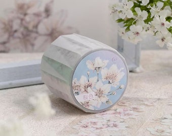 Puede incluir: Un rollo de cinta adhesiva decorativa con un diseño floral. La cinta presenta una combinación de colores azul claro y blanco con un patrón repetido de flores de cerezo blancas. El texto "桜色" está impreso en la cinta.