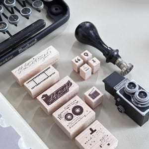 May include: A collection of wooden rubber stamps featuring various designs including a film strip, the text Life is beautiful, a cassette tape with the text Record, and small icons. A black wax seal stamp and a miniature black Ricohflex camera are included.