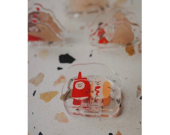 May include: Clear acrylic charm depicting a smiling ketchup bottle and a hot dog, holding hands. The charm has a transparent background with a white and red design. Other similar charms are visible in the background.