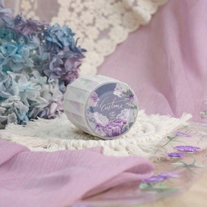 May include: A roll of decorative washi tape with a floral design in purple and white. The tape is labelled "Customa" in cursive. The tape is placed on a white macrame coaster, with a soft purple fabric in the background.