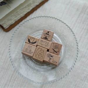 Puede incluir: Un conjunto de ocho sellos de madera con varios diseños, incluyendo un cisne, una flor, una ardilla, un mapache, un libro, un pastel y un pájaro. Los sellos están dispuestos en un círculo sobre un plato de vidrio transparente.