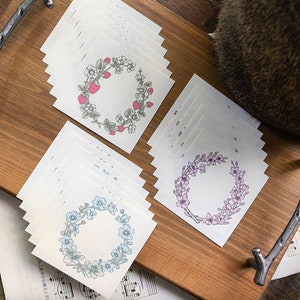 May include: A set of 12 blank cards featuring floral wreath designs. The wreaths are in three different colours: pink, blue, and red. The cards are ideal for journaling, scrapbooking, or card making.