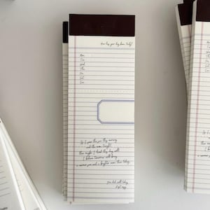 May include: A vertical notepad with a dark brown header and white paper featuring blue horizontal lines and red vertical margins. The page includes a weekly tracker, a blank rectangular box, and printed cursive text that reads, How has your day been truly? and You did well today.