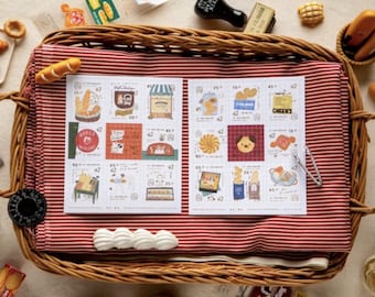 May include: A wicker basket with a red and white striped cloth contains two sheets of illustrated stickers with bakery designs. Miniature breads and pastries surround the basket, creating a charming food-related display.