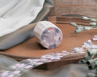 Puede incluir: Un rollo de cinta adhesiva decorativa washi con un diseño floral. La cinta presenta un fondo morado claro con flores rosas y blancas y hojas verdes. La cinta está sobre una superficie marrón con una tira de cinta floral a juego.