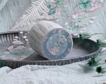 Puede incluir: Un rollo de cinta adhesiva decorativa washi con un diseño floral. La cinta presenta un fondo verde azulado con flores rosas y blancas. El embalaje es blanco y translúcido, y la cinta se muestra en una bandeja gris.