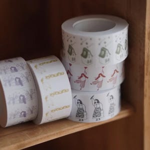 May include: Several rolls of decorative washi tape with various patterns. One roll features a repeating design of women, another has a floral pattern, and others have houses and figures. The tape is displayed on a wooden shelf.