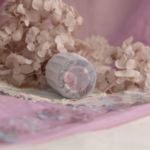 May include: A decorative roll of washi tape with a pink rose design. The tape is wrapped in white, textured paper and is set against dried flowers and pink fabric. The label on the tape features Chinese characters.