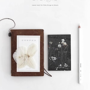 May include: A brown leather-bound notebook featuring a pressed white flower on the cover, a rectangular card with a dark floral print, and a white pencil. The text reads Memopad, 35 / 36 / 37 / 38, Leave room for little things to bloom, and NYRET DESIGN.