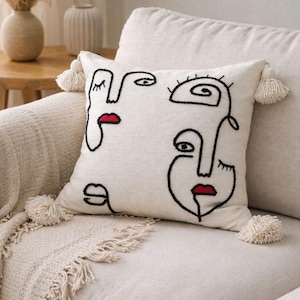 May include: Cream cushion featuring embroidered abstract faces in black thread and red lips. The cushion has white tassels on each corner and is on a beige sofa. Home decor item.