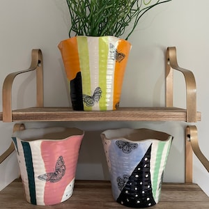 May include: Three ceramic planters with geometric patterns and butterfly designs. The planters are on a wooden shelf with gold metal brackets.