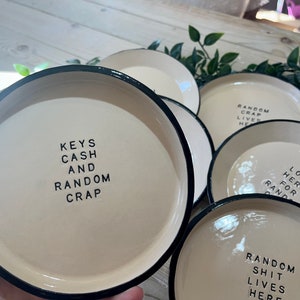 Może przedstawiać: Pięć ceramicznych miseczek z czarnymi obw&oacute;dkami i białym wnętrzem. Każda miseczka ma inny napis wydrukowany czarnym kolorem. Napisy to: "Keys Cash and Random Crap", "Random Crap Lives Here", "Look Here For Random" i "Random Shit Lives Here".