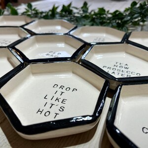 Puede incluir: Un juego de seis platos hexagonales de cer&aacute;mica blanca con bordes negros. Cada plato tiene un dicho diferente, incluyendo "Drop it like it's hot", "Too hot to hold", "Tea now Prosecco later", y "Old Bag".