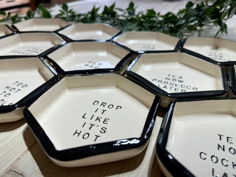 Puede incluir: Un juego de seis platos hexagonales de cer&aacute;mica blanca con bordes negros. Cada plato tiene un dicho diferente, incluyendo "Drop it like it's hot", "Too hot to hold", "Tea now Prosecco later", y "Old Bag".