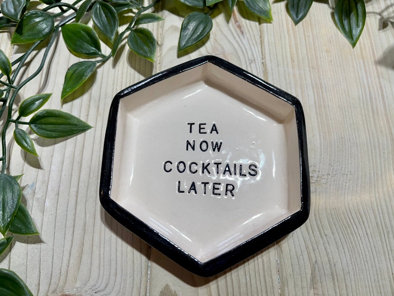 Puede incluir: Un plato hexagonal de cer&aacute;mica blanca con un borde negro. El plato tiene el texto "TEA NOW COCKTAILS LATER" escrito en negro.