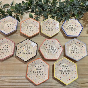 Puede incluir: Conjunto de diez platos hexagonales de cer&aacute;mica con lunares coloridos y refranes humor&iacute;sticos. Los refranes incluyen "T&eacute; ahora, vino despu&eacute;s", "Demasiado caliente para manejar", "T&eacute; ahora, c&oacute;cteles despu&eacute;s", "T&eacute; ahora, ginebra despu&eacute;s", "T&eacute; ahora, burbujas despu&eacute;s", "T&eacute; ahora, prosecco despu&eacute;s", "T&eacute; ahora, estrellas porno despu&eacute;s", "Bolsa de t&eacute; yo", "Derrama el t&eacute;" y "Pon la bolsa aqu&iacute;".