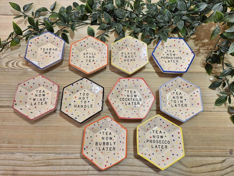 Puede incluir: Conjunto de diez platos hexagonales de cer&aacute;mica con lunares coloridos y refranes humor&iacute;sticos. Los refranes incluyen "T&eacute; ahora, vino despu&eacute;s", "Demasiado caliente para manejar", "T&eacute; ahora, c&oacute;cteles despu&eacute;s", "T&eacute; ahora, ginebra despu&eacute;s", "T&eacute; ahora, burbujas despu&eacute;s", "T&eacute; ahora, prosecco despu&eacute;s", "T&eacute; ahora, estrellas porno despu&eacute;s", "Bolsa de t&eacute; yo", "Derrama el t&eacute;" y "Pon la bolsa aqu&iacute;".