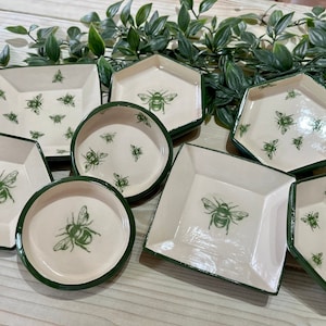 May include: Set of seven ceramic dishes with green trim and bee designs. The dishes are in various shapes, including square, round, and hexagonal.