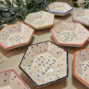 Puede incluir: Un juego de nueve platos hexagonales de cer&aacute;mica con lunares coloridos y refranes humor&iacute;sticos. Los refranes incluyen "Spill the Tea", "Place Bag Here", "Too Hot to Handle", "Tea Now Cocktails Later", "Tea Now Gin Later", y "Tea Now Bubbly Later".