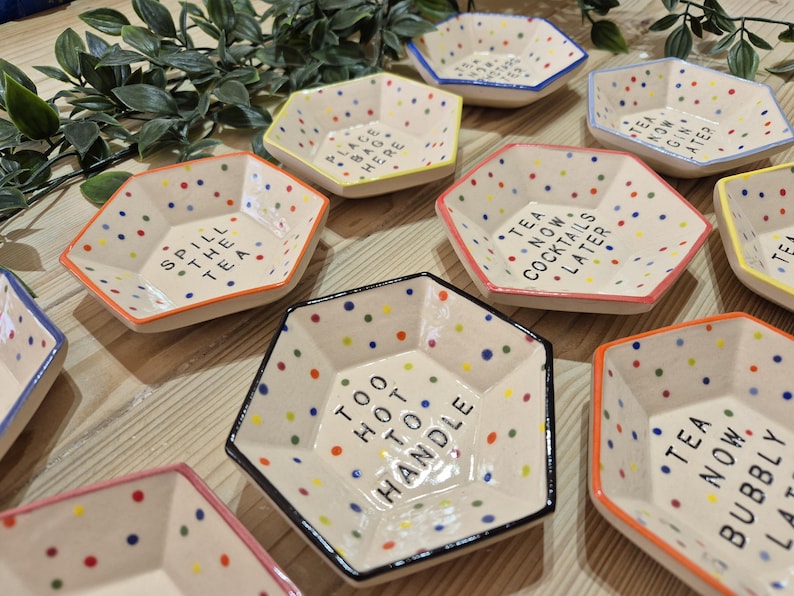 Puede incluir: Un juego de nueve platos hexagonales de cer&aacute;mica con lunares coloridos y refranes humor&iacute;sticos. Los refranes incluyen "Spill the Tea", "Place Bag Here", "Too Hot to Handle", "Tea Now Cocktails Later", "Tea Now Gin Later", y "Tea Now Bubbly Later".